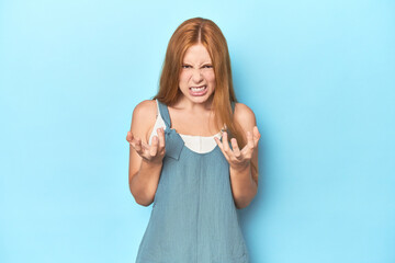 Redhead young woman on blue background upset screaming with tense hands.