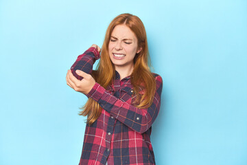 Redhead young woman on blue background massaging elbow, suffering after a bad movement.