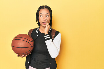 Indonesian schoolgirl with basketball on yellow is saying a secret hot braking news and looking...