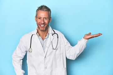 Caucasian middle-aged doctor on blue background showing a copy space on a palm and holding another...