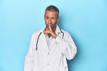 Caucasian middle-aged doctor on blue background keeping a secret or asking for silence.