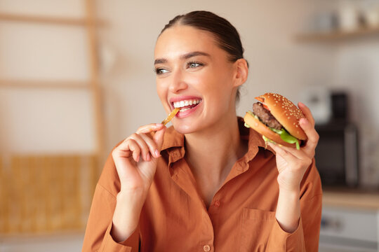 Fast food lover. Happy lady eating burger and french fries, biting chips and looking aside, sitting in kitchen at home