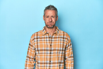 Fototapeta premium Middle-aged caucasian man on blue backdrop blows cheeks, has tired expression. Facial expression concept.