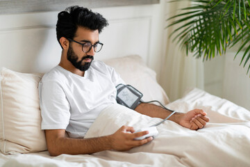 Indian Man Using Upper Arm Blood Pressure Monitor While Sitting In Bed