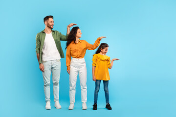 Fototapeta premium Family hierarchy. Young parents and daughter measuring each other's height with hand, posing over blue background