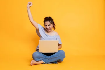 Zelfklevend Fotobehang Portrait of attractive cheerful girl sitting in lotus pose using laptop on isolated over bright background, Female excited with offers and winning over internet © Photographielove