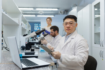 International group of young scientists work in the laboratory. An Asian man is sitting at a desk and using a tablet, behind which colleagues are examining with a microscope.