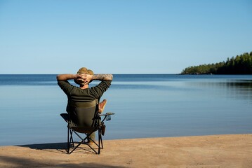 Man rests on a camping chair and looks out over the lake on a sunny day - obrazy, fototapety, plakaty