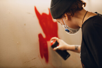 Young woman with a spray balloon with paint in her hand in a protective mask painting wall garage...
