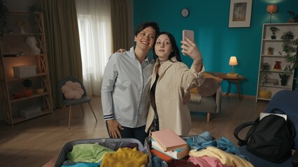 Photo of a teenager, young woman having a videocall, making a video, taking a photo, selfie with her mother helping her daughter to pack her belongings in a suitcase.