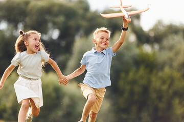 Holding each other by hands. Boy and girl are playing with toy plane on the green field