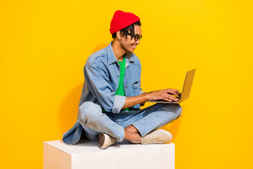 Full body photo of attractive young man sitting white platform netbook typing wear trendy jeans clothes isolated on yellow color background