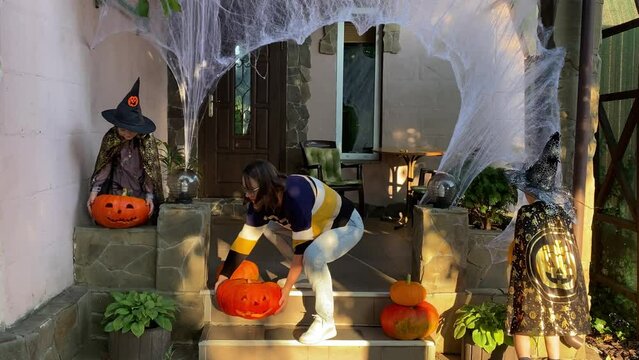 Family enthusiastically prepares for Halloween outdoors. A mother and kids joyfully decorate the front porch with Jack-o'-lanterns, creating a festive atmosphere filled with laughter and teamwork