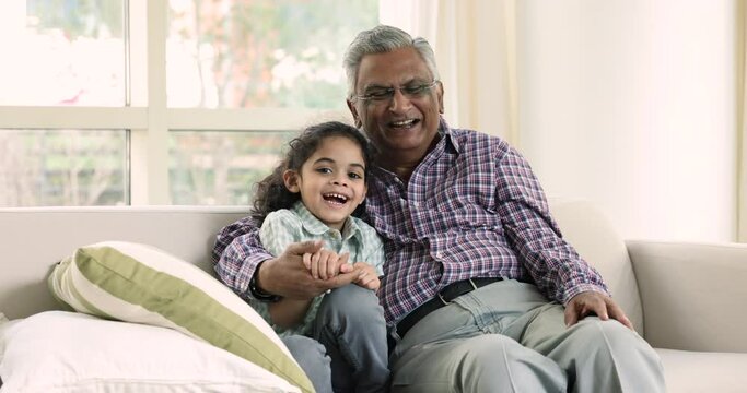 Loving elderly man hugging his little cute great-grandson. Preschooler boy posing for camera seated on sofa at home with caring mature great-grandfather. Indian multigenerational family ties and bond
