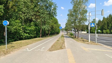 Fototapeta na wymiar Tile-paved bike and pedestrian paths, marked with signs and pavement markings, are located between the park's trees and the roadway of the city street where cars travel. Summer sunny weather