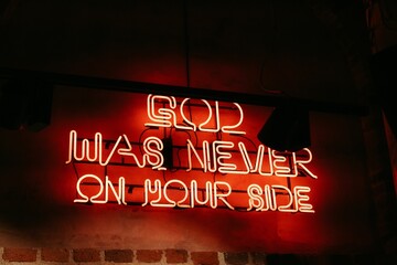 Brightly lit neon sign with the phrase "God Was Never On Our Side" in a vivid font
