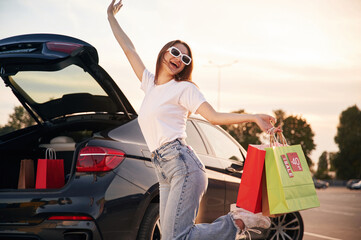 Against the car with trunk opened. Beautiful woman in casual clothes is holding shopping bags, outdoors