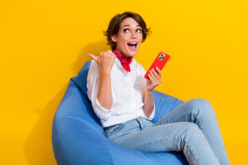 Photo of cheerful surprised girl sitting comfort pouf hold iphone demonstrate wow offer empty space...