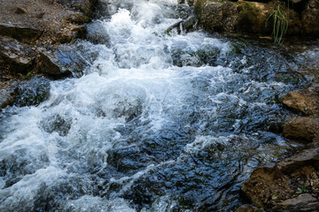 A stormy stream of a mountain river flows over large and small stones, white waves, strong current, small waterfalls, swirls, dark water