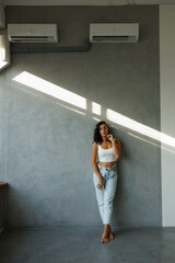 Beautiful brunette woman in white top and blue jeans, standing. Girl smile, happy. Portrait of young pretty woman. Complicated sunlight with shadows. White background. Light from window, portrait.