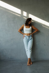 Beautiful brunette woman in white top and blue jeans, standing. Girl smile, happy. Portrait of young pretty woman. Complicated sunlight with shadows. White background. Light from window, portrait.