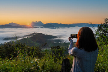 Fototapeta na wymiar Female tourists backpacks, travel, hike, hold smartphones to take pictures of the scenery and admire the sea of mist from the mountain top and feeling free, good weather in the morning, weekend 