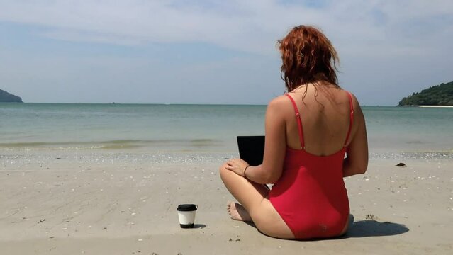 Young woman wearing orange swimsuite working on the beach with laptop and drinking coffee