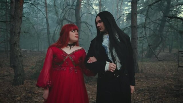 Halloween couple costume vampire old style gothic fantasy man and woman walking in deep dark forest tree. king in black tailcoat long hair, guy holding hand of girl Queen. Look at each other love 4k