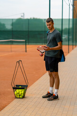 young professional player coach standing on outdoor tennis court before starting game training with racket and basket of tennis balls