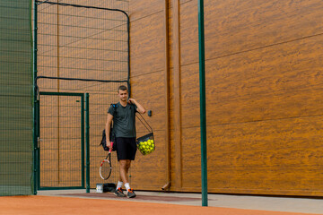 young professional player coach standing on outdoor tennis court before starting game training with racket and basket of tennis balls