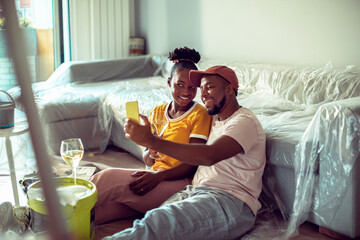 Young African American couple taking a selfie on smartphone while drinking wine on a break from painting their living room at home