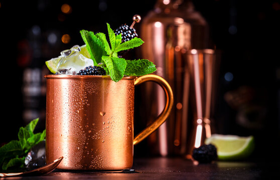 Moscow mule cocktail drink with blackberry in copper mug with lime, ice, ginger beer, vodka and mint. Black bar counter background, bar tools, bottles