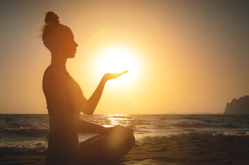 Fototapeta premium Side view. Silhouette shot at sunset or sunrise. Young caucasian woman meditating in the lotus position on the seashore holds the sun on her hand. Yoga and meditation practice at sunrise by the sea