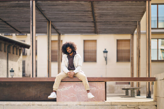 a young man with afro hair sitting on a high step in the square with his legs dangling down