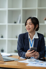 Asian businesswoman using mobile phone to communicate, chat, negotiate, search internet, news, email while working online on laptop computer at office.