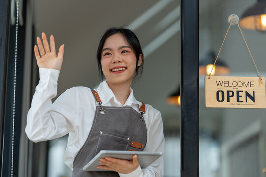 Young freelance businesswoman Asian young female barista in apron holding sign preparing to open for sale and standing in front of the door of the cafe with open sign business owner startup concept