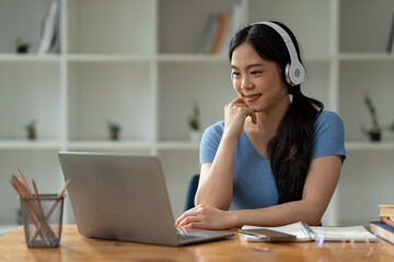 Asian woman wearing headphones sitting and working Listen to relaxing music and chat with friends on social media on laptop while sitting on weekends home office desk.