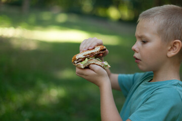 the cute little boy examines with interest the juicy hamburger he has just bitten off. wearing a...