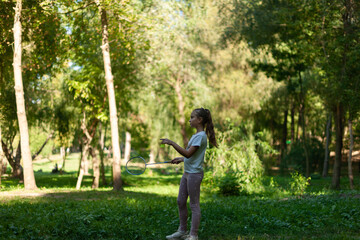 Obraz na płótnie Canvas little cute caucasian girl, playing badminton in a summer park, surrounded by green trees