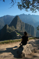 Young woman with long flowing hair, wearing sportswear, sitting on a rock watching the archeological site of Machupicchu, Peru. Views of the mountains, with a clear and sunny sky. 
