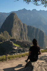 Young woman with long flowing hair, wearing sportswear, sitting on a rock watching the archeological site of Machupicchu, Peru. Views of the mountains, with a clear and sunny sky. 