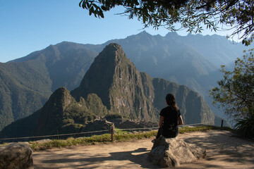 Young woman with long flowing hair, wearing sportswear, sitting on a rock watching the archeological site of Machupicchu, Peru. Views of the mountains, with a clear and sunny sky. 