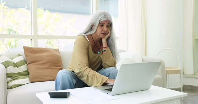 Aged woman sit on sofa use laptop and calculator manage budget, feels upset due to lack of money to pay domestic utilities, having unpaid bills, high taxes. Subpoena, financial problem of older female