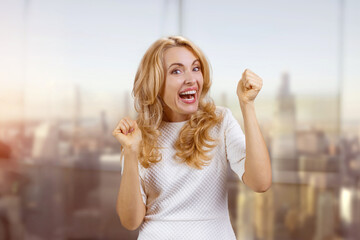 Happy excited blonde woman rejoicing something in the office. Blurred evening cityscape in the background.