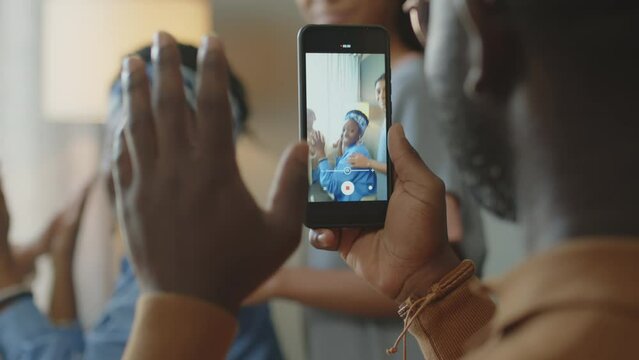 Selective focus shot of black man holding mobile phone and filming video of cheerful wife and daughters playing patty cake and waving during holiday dinner at home