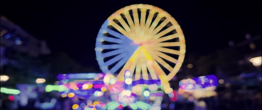Illuminated Ferris wheel and carousels in the evening at the Brilon fair. The lights and movements can be seen as blurred bokeh. Unknown visitors stroll by. Real-time video in anamorphic format 1.33.