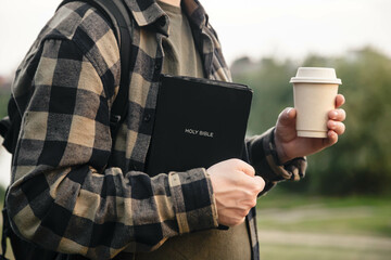 Bible and paper cup in the hands of a Christian man outside on a blurred background of nature.