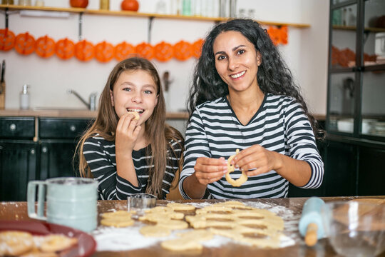 Portrait of two people, mother and daughter sitting in the kitchen at the table baking cookies from dough. Oriental brunette woman and Caucasian girl trying Playing with food smiling lookin at camera