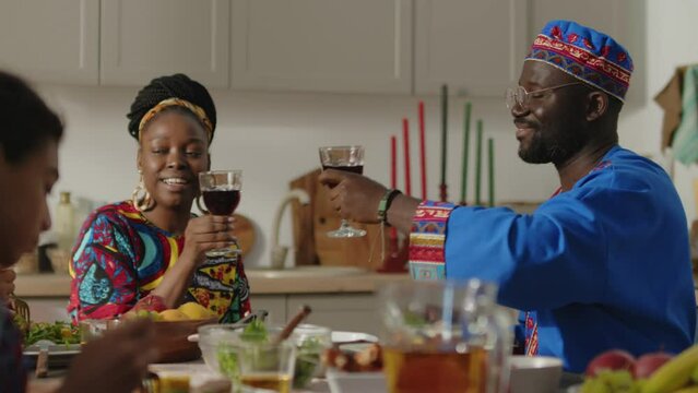 Cheerful African American wife and husband in traditional dashiki tunics clinking glasses in toast, drinking red wine and having talk with kids on Kwanzaa dinner at home