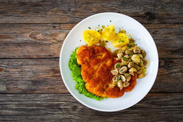 Crispy breaded seared chicken cutlet with fried white mushrooms and boiled potatoes on wooden table
